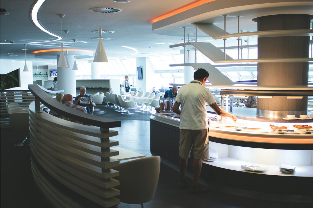 Passengers sit at the airport lounge enjoying a meal. A man in shorts picks food from the buffet. 