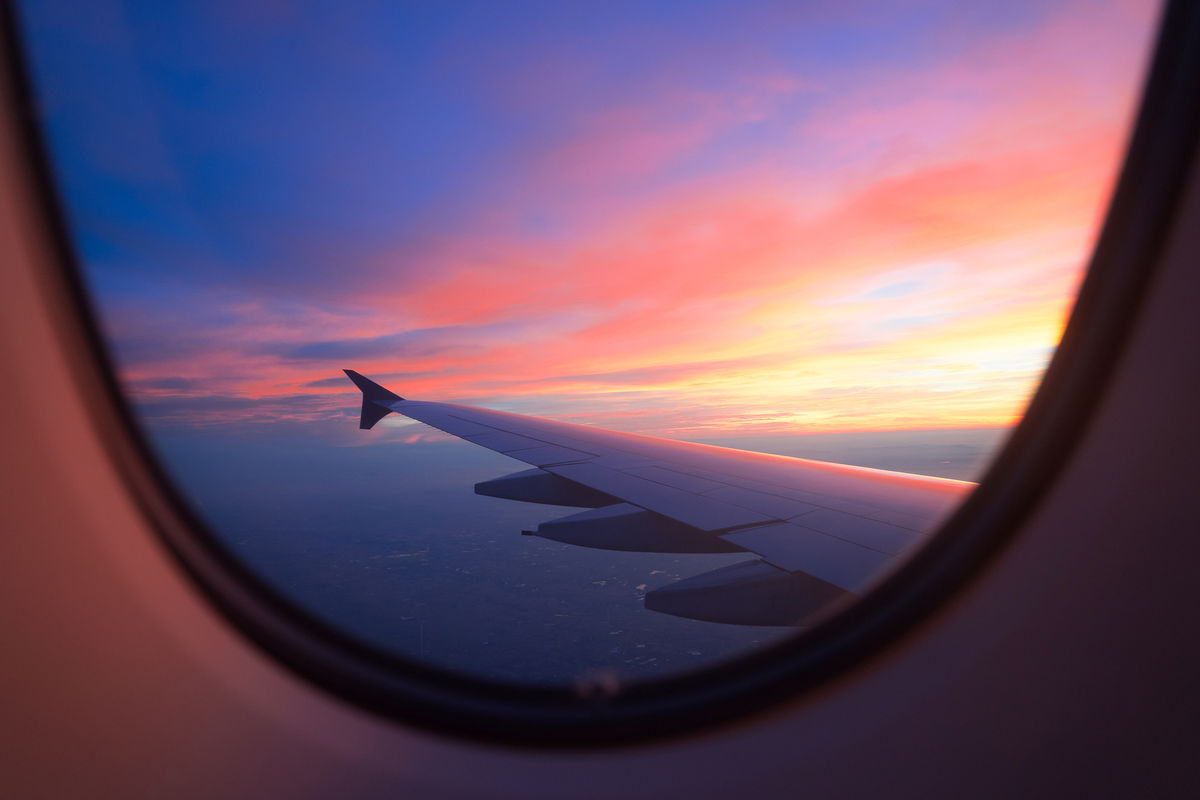 A view from an airplane window showing the wing illuminated by the warm hues of a vibrant sunset. The sky transitions from deep blue to shades of pink, orange, and purple, creating a serene and colorful scene.