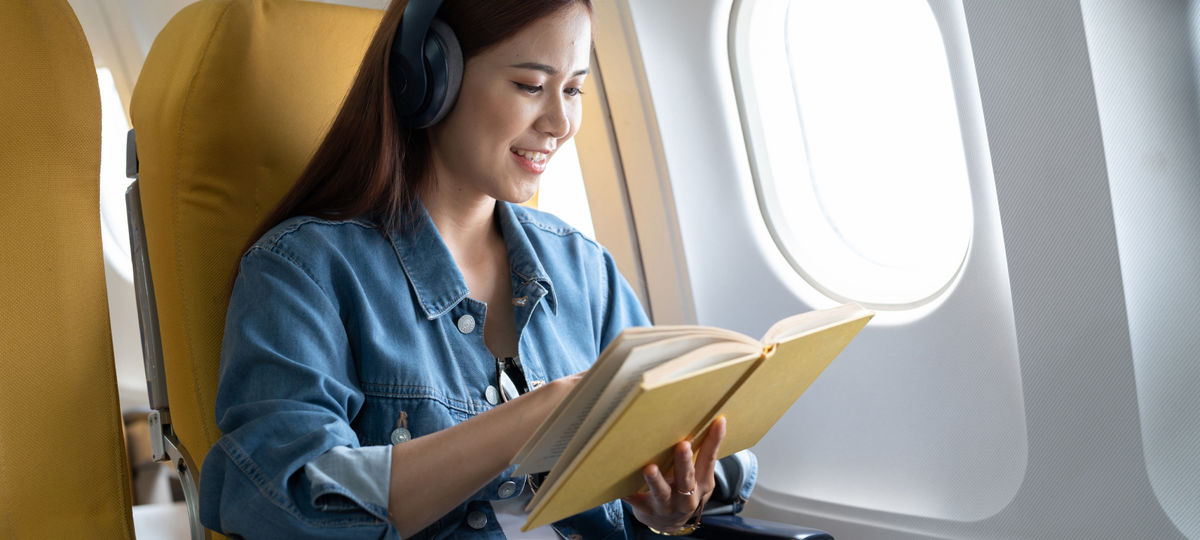 A woman is reading a book on a plane while wearing headphones.