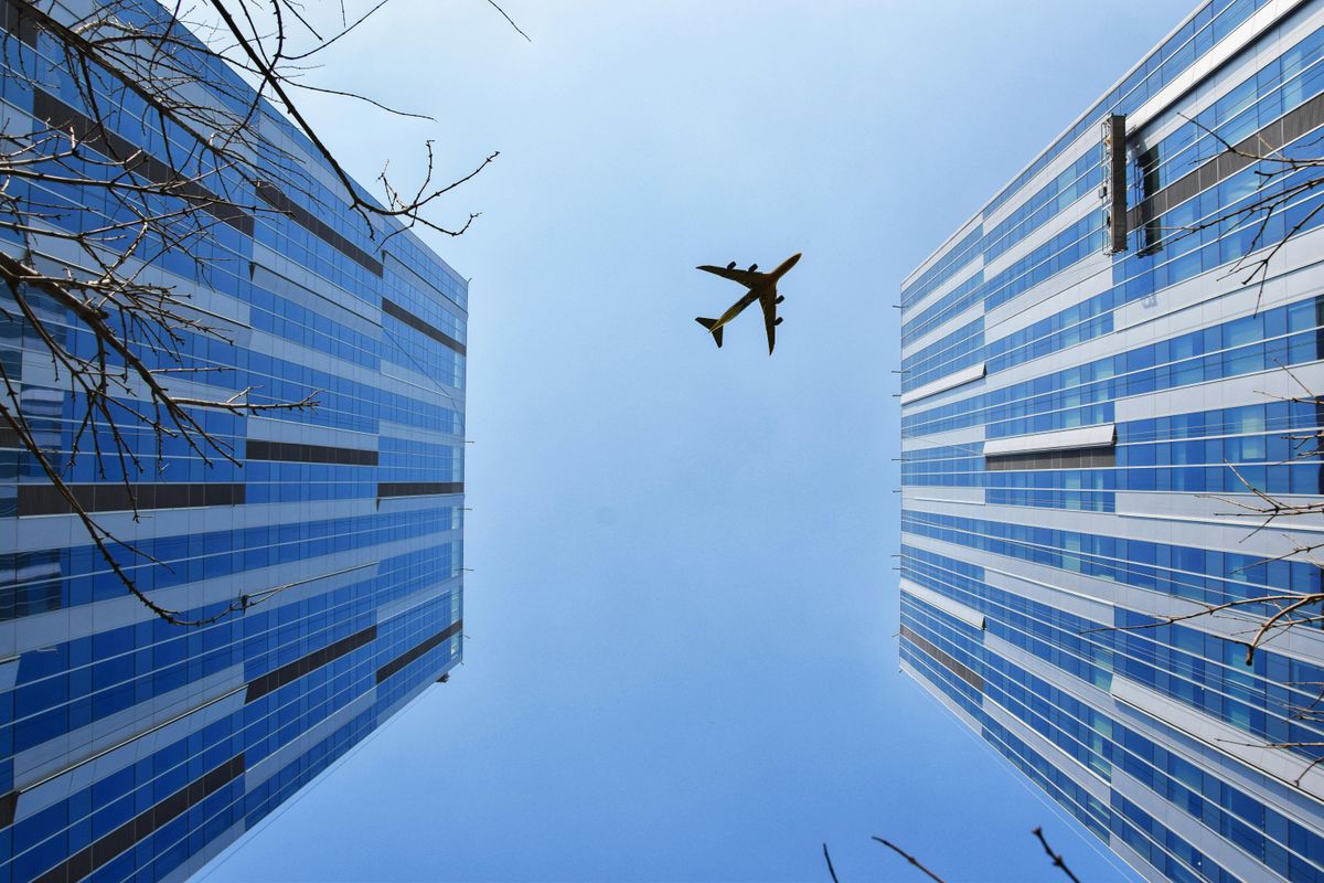 An airplane flying overhead, captured between two modern glass skyscrapers under a clear blue sky. The perspective looks upward, with bare tree branches adding a natural element to the urban scene.