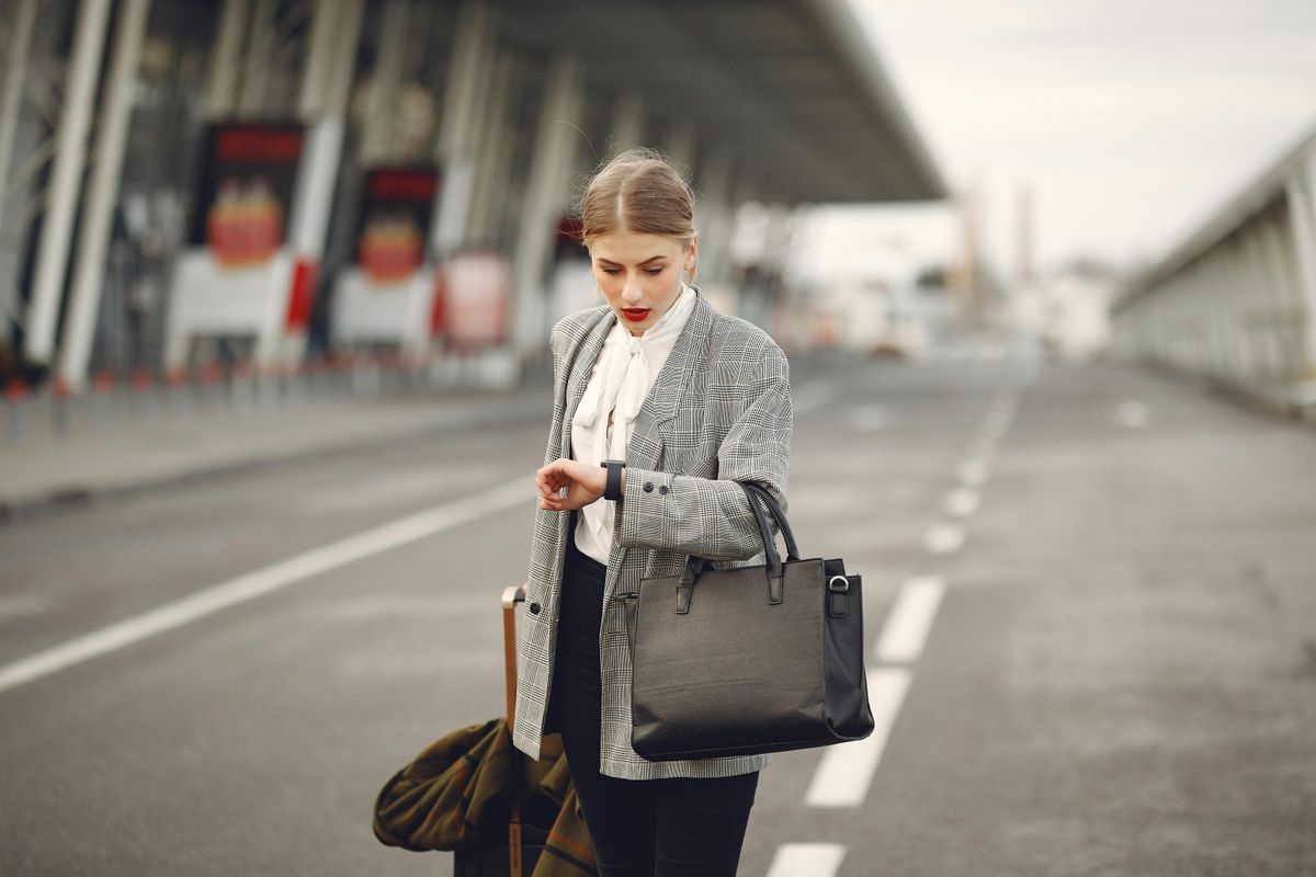 A passenger, wearing a smart, grey blazer checks her watch outside the airport with a hurried expression on her face. 