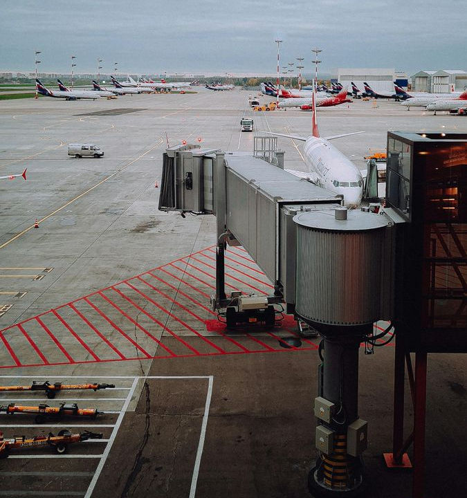 A photo of a jet bridge extending out from the airport terminal building. There are numerous planes on the tarmac in the distance.