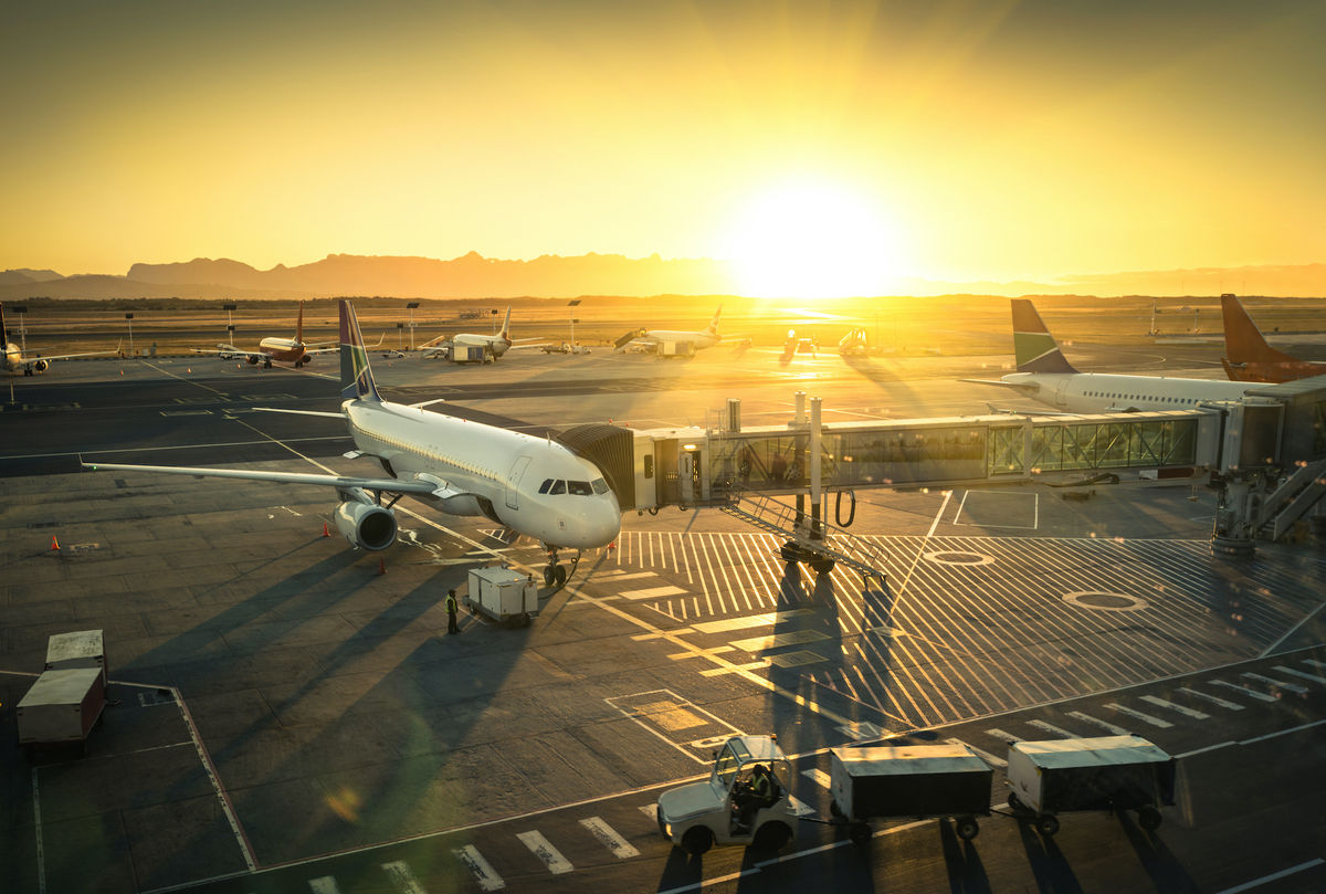 A white airplane parked at the gate with a jet bridge attached as passengers board. The setting sun casts a warm yellow and orange glow across the airport, where other planes are visible in the distance. The silhouette of jagged hills frames the horizon, adding depth to the scene.