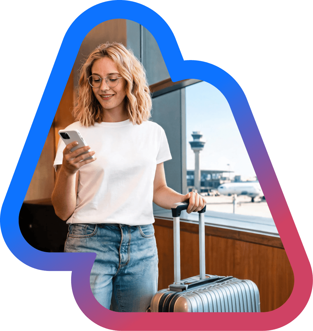 A smiling young woman using her smartphone while standing with a suitcase at an airport terminal