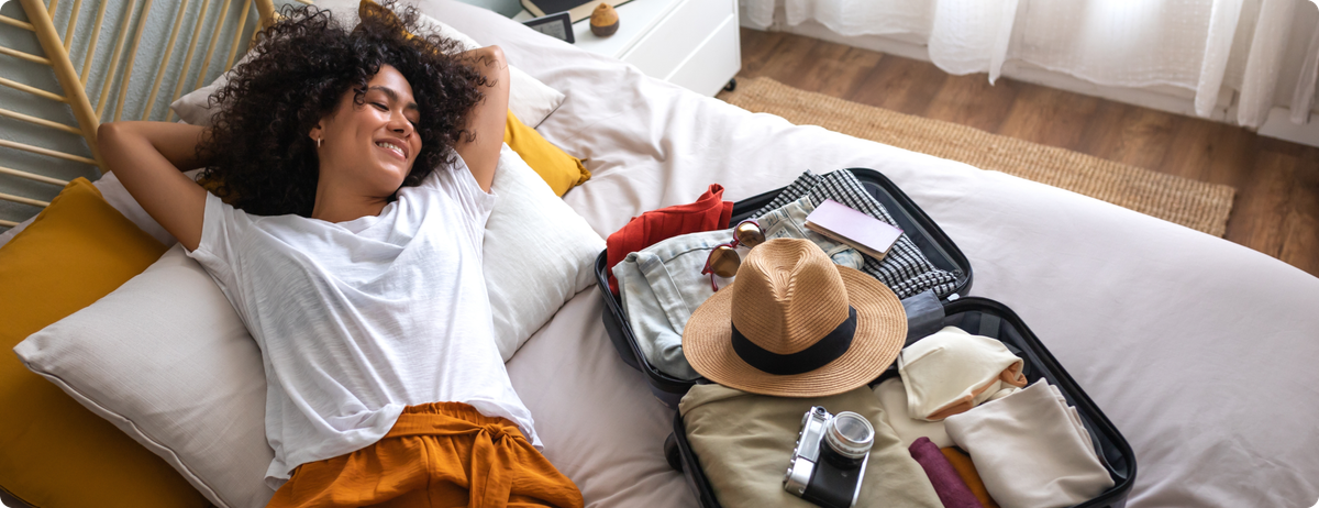 A woman relaxes on her bed next to her full suitcase of luggage