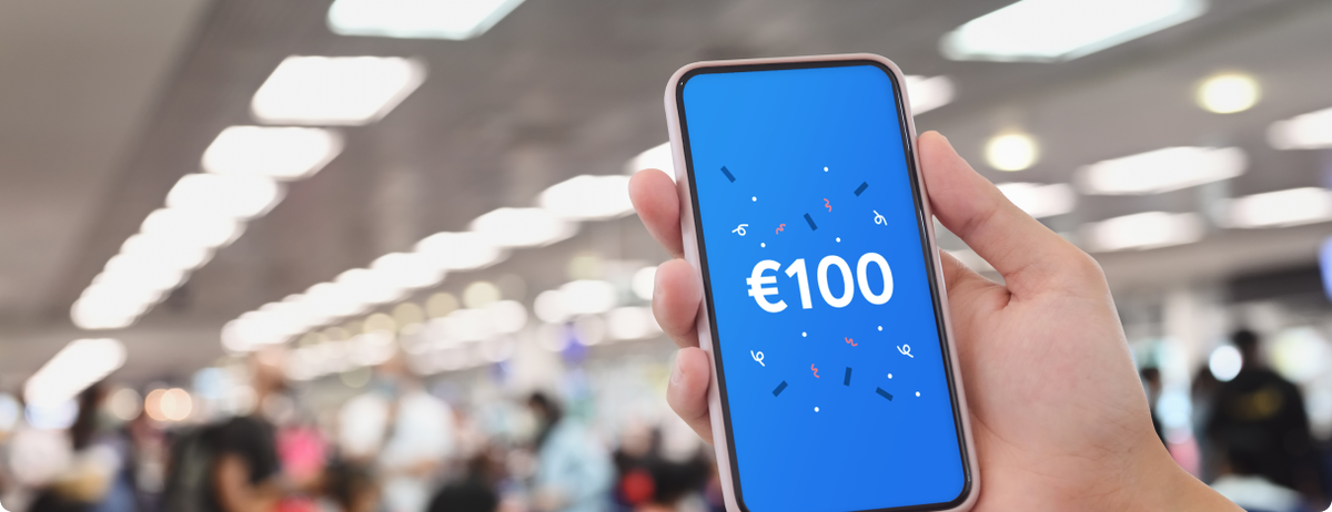 Hand holding a mobile phone showing a €100 payout from AirPayout insurance. the blurred background shows this passenger is in a busy airport